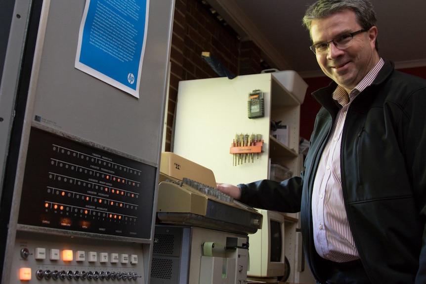A man stands next to a large grey box, lined with lights and switches, attached to a teletype machine.