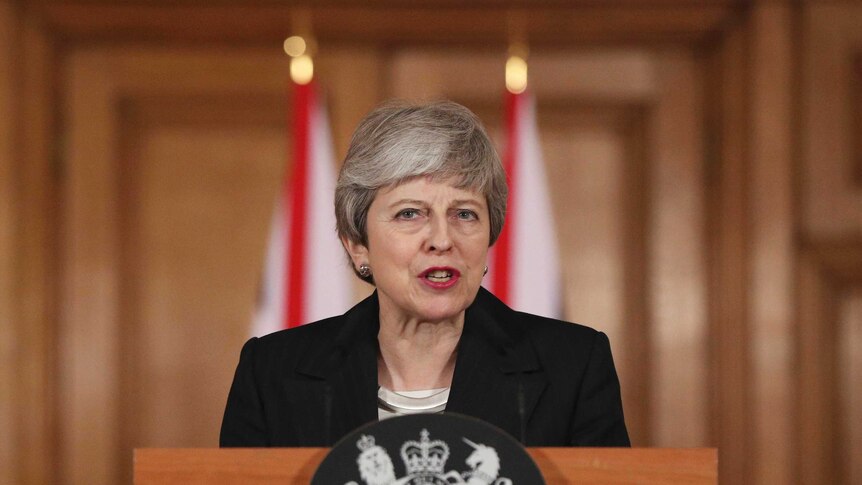 Theresa May speaks in front of a warm wooden lectern with British insignia in front of two union jack flags and wooden panels.