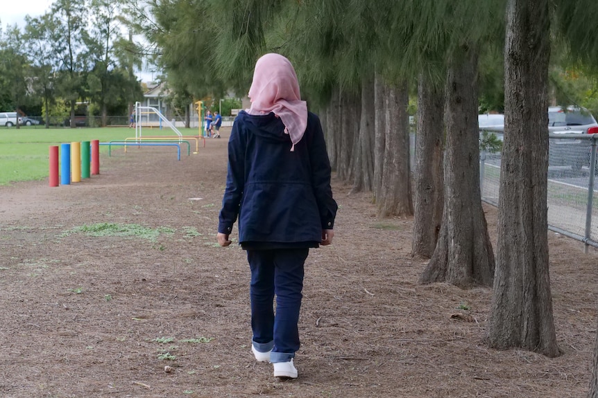 A teenage girl wearing a pink hijab, jeans and a navy blue jacket walks in a park with her back to the camera.