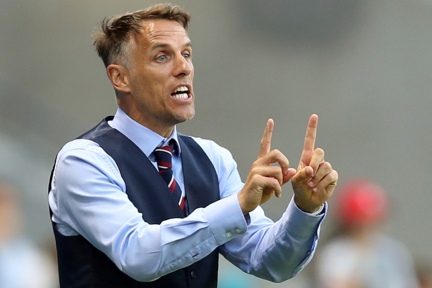 Phil Neville stands in a waistcoat holding one finger up on each hand, holding the fingers parallel to each other