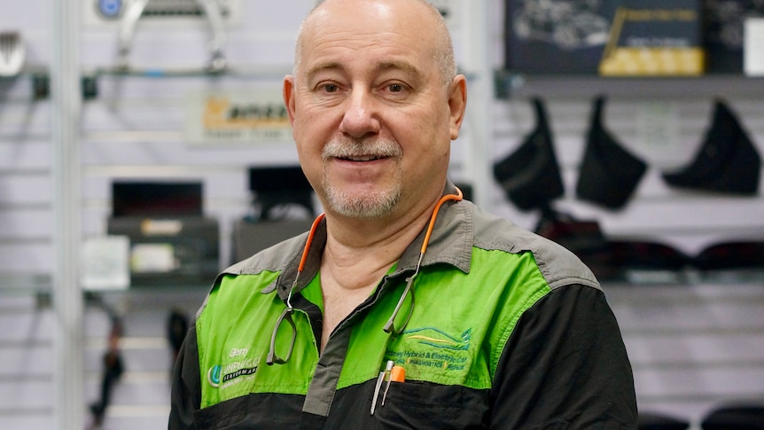A bald man wearing a black and green mechanic shirt stares at the camera.
