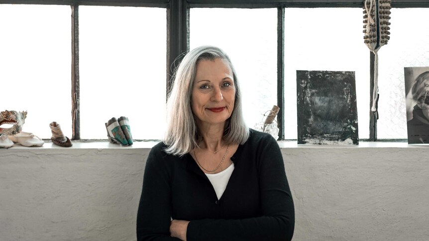 The artist Julie Rrap smiles at the camera. She has long grey hair and is crossing her arms.