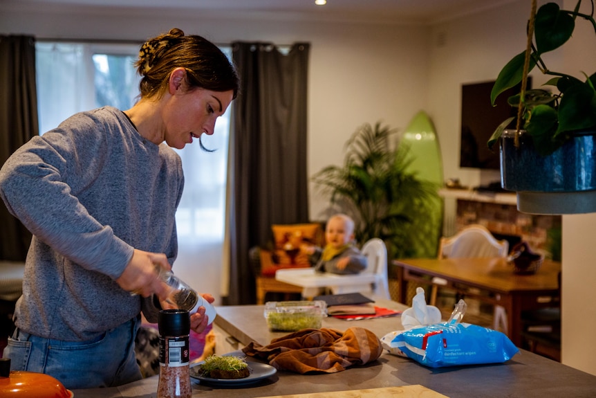 Ginelle cracks pepper onto a meal in the kitchen with her baby in a high chair behind her.