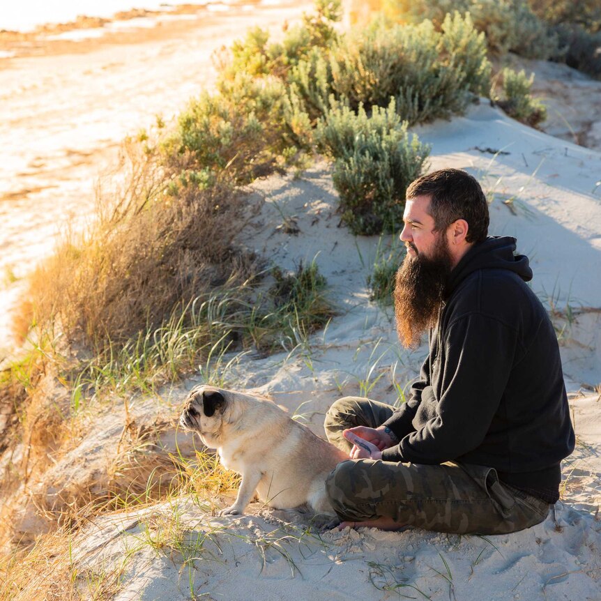 A young bearded man and his pug sit on a sand dune staring out to sea on a bright sunny day.