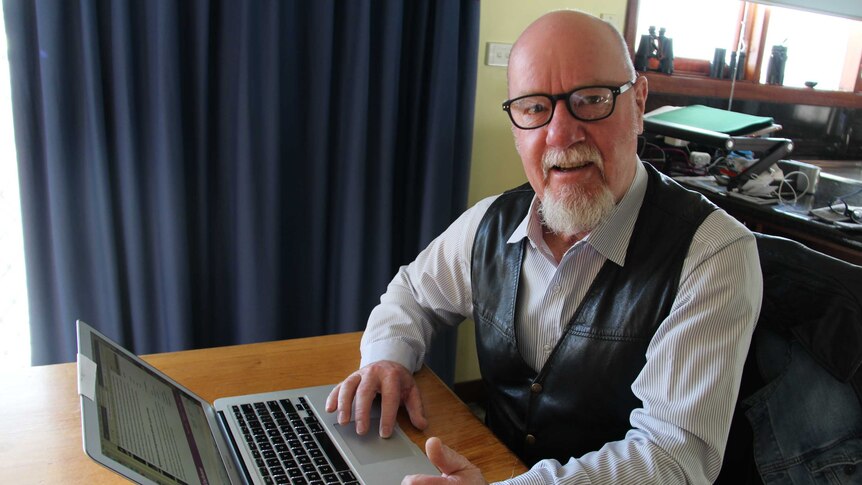A man in a striped shirt and leather vest sits in front of a laptop at at table.