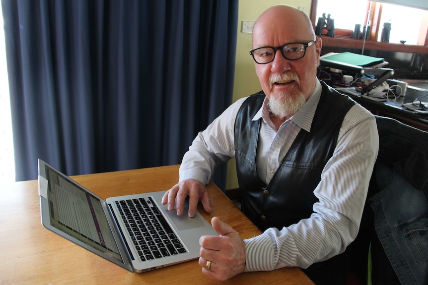 A man in a striped shirt and leather vest sits in front of a laptop at at table.