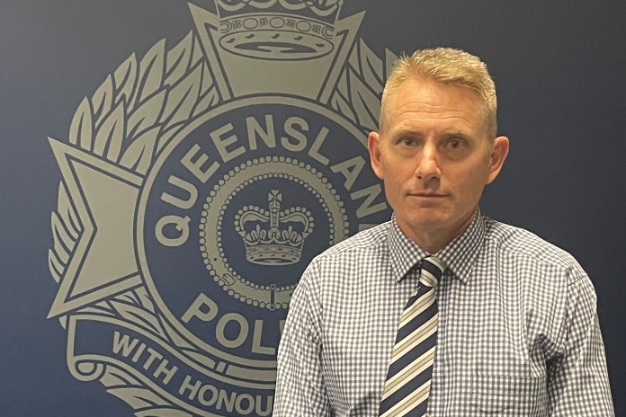 A senior detective standing in front of the Queensland Police Service logo
