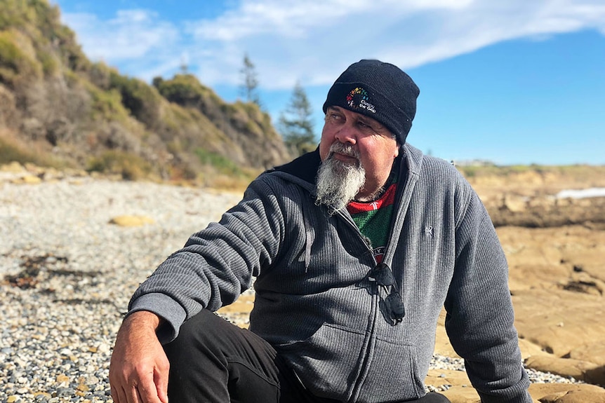An older man wearing a black beanie, grey jumper and black jeans is also sporting a grey, narrow beard as he sits on rocks