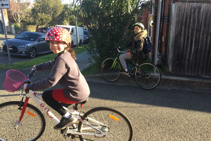 Two young girls ride bicycles down a suburban street.