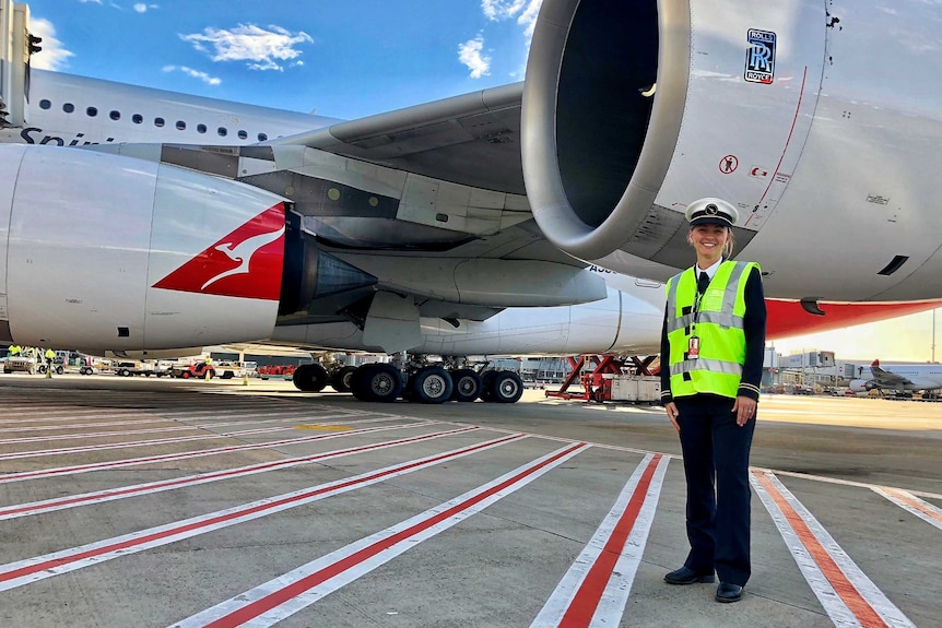 A female pilot in a high-vis vest stands next to an engine of a Qantas passenger jet at an airport.