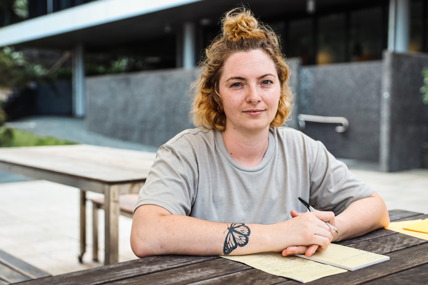 A woman looks to the camera as she leans over a notepad on a table