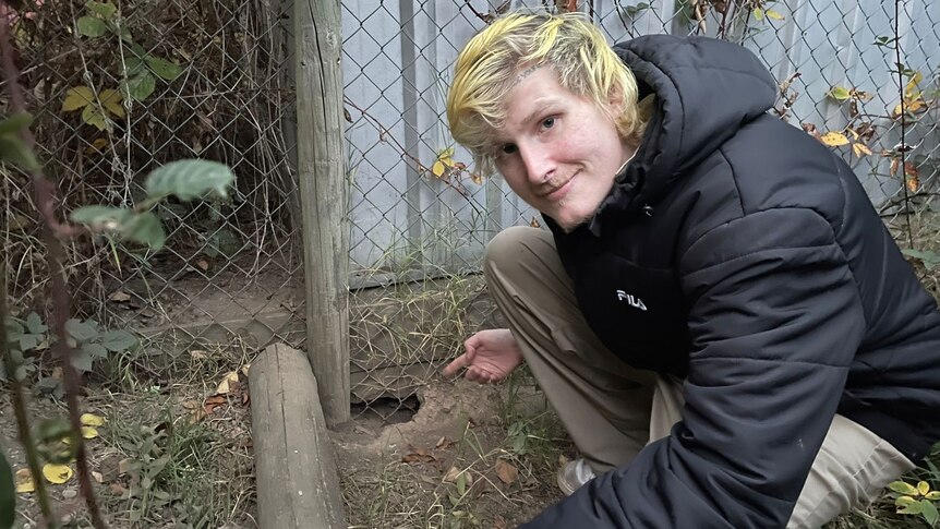 man with blonde hair smiling and crouching in a garden bed pointing to hole in theh ground