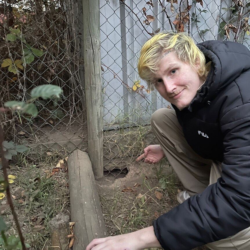 man with blonde hair smiling and crouching in a garden bed pointing to hole in theh ground