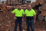 Two men in high-visibility shirts stand facing the camera with piles of logs behind them.
