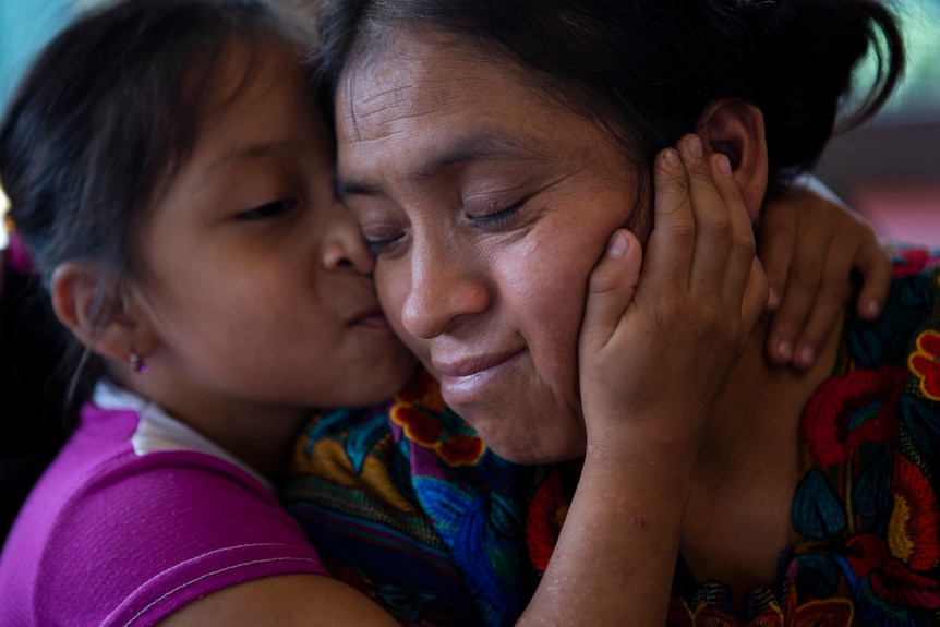 A young guatemalen girl with dark hair and features holds her mother's face close to hers and kisses her, both shut their eyes 