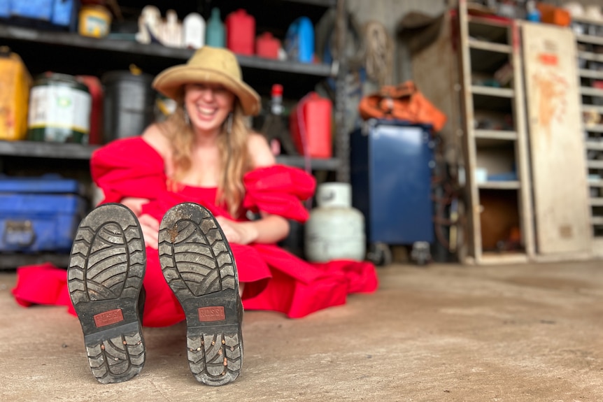 A woman wearing a red dress, hat and boots, sitting in front of a garage shelf, laughing.