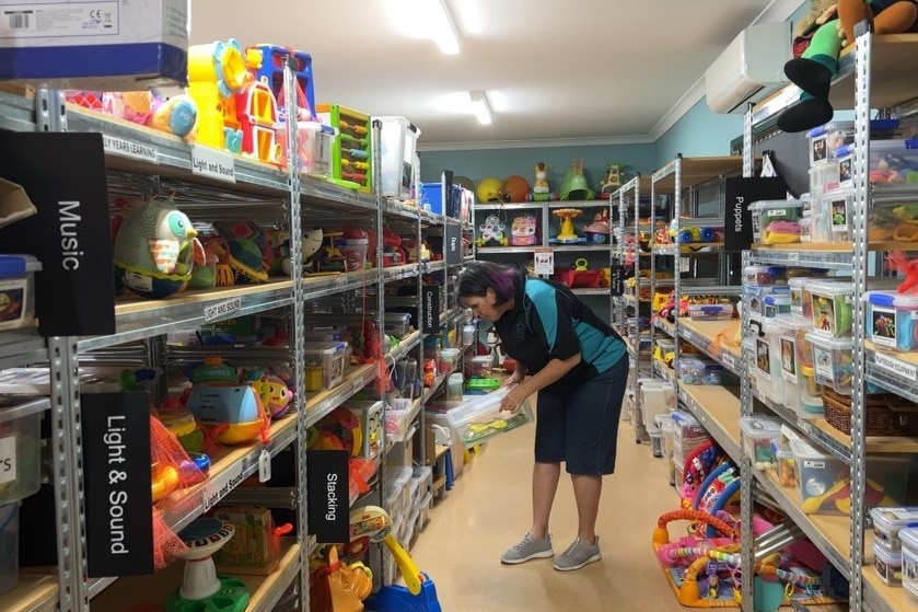 A woman in a polo shirt and jean shorts bends over, she is surrounded by shelves of toys