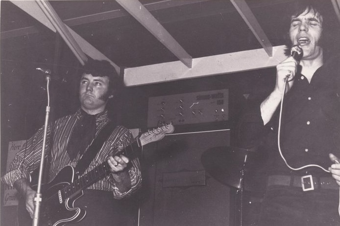 A black and white photo two men with big sideburns, one playing a guitar, the other singing into a mic with amp in background.