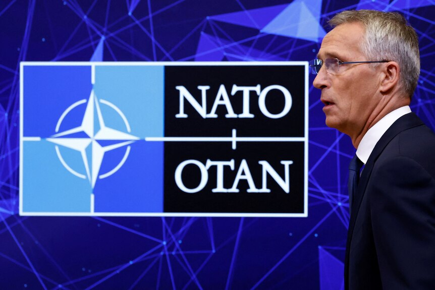 Jens Stoltenberg photographed on the side, in a suit, speaking in front of a screen with 'NATO OTAN' on it