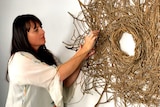 A woman with long dark hair stands near a sculpture which uses branches and sticks to represent the sun.