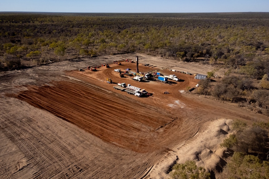 An aerial view of several lines of trucks parked on a large, cleared patch of dirt, surrounded by scrub.