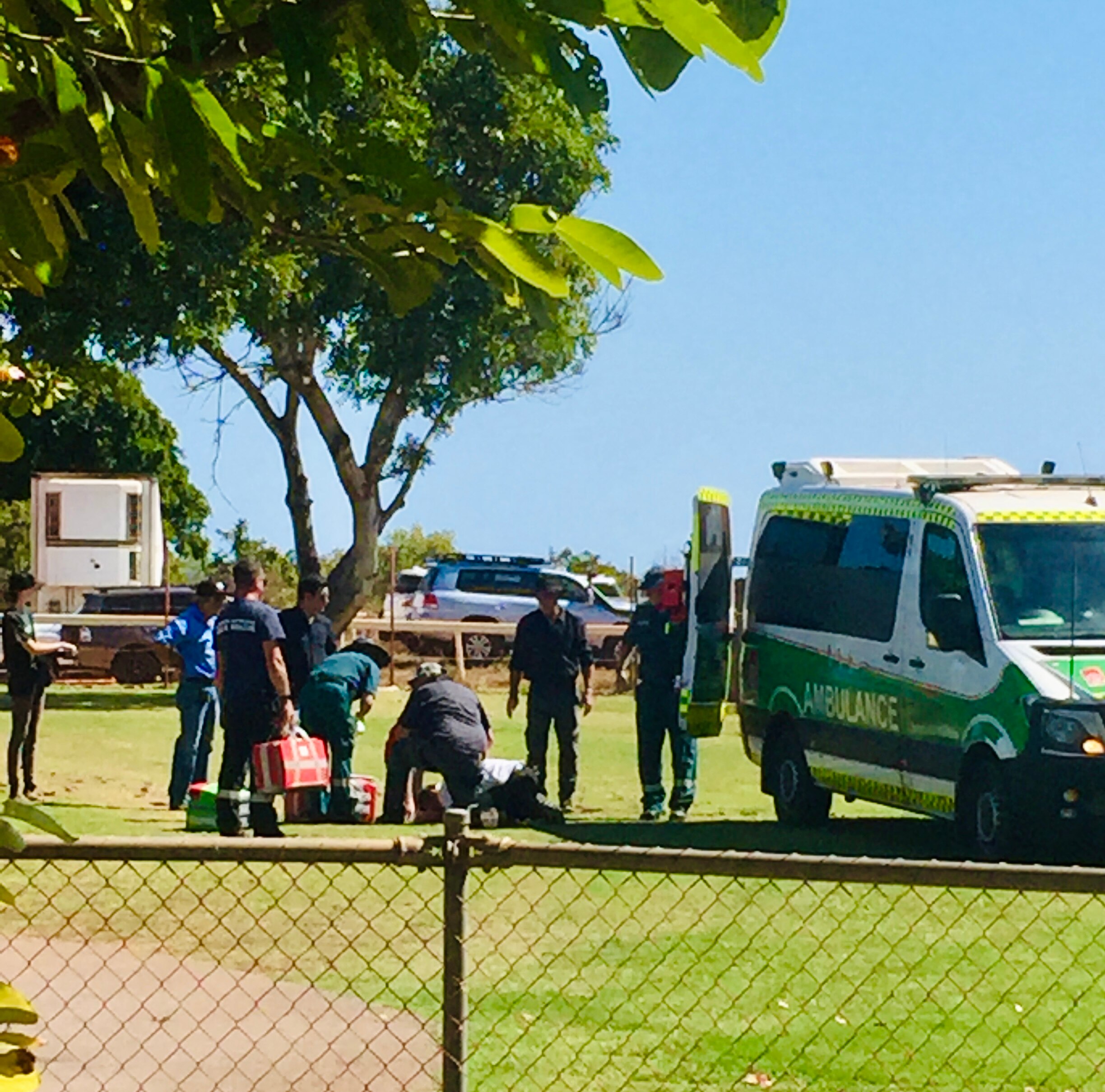 Paramedics gather around a person on the ground in a park with an ambulance parked nearby