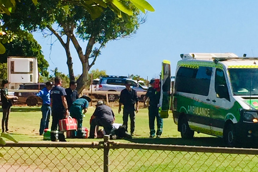 Paramedics gather around a person on the ground in a park with an ambulance parked nearby