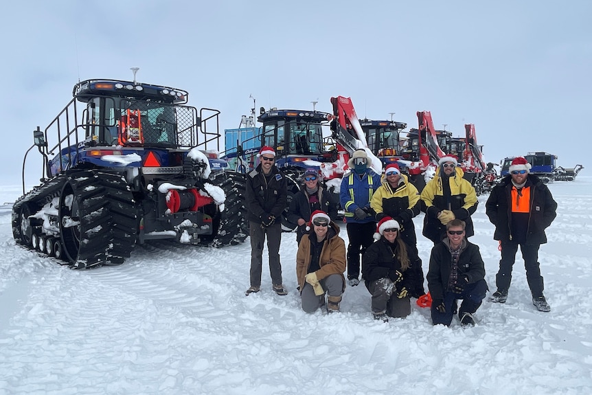 A group of people wearing Santa hats stand in front of a line of vehicles in the snow.