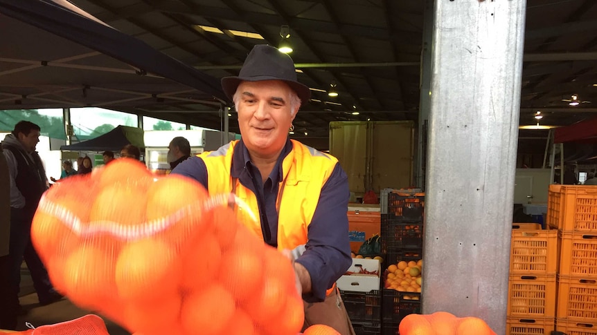 Man in orange vest throws large bag of oranges on a table. He wears a hat and a smile