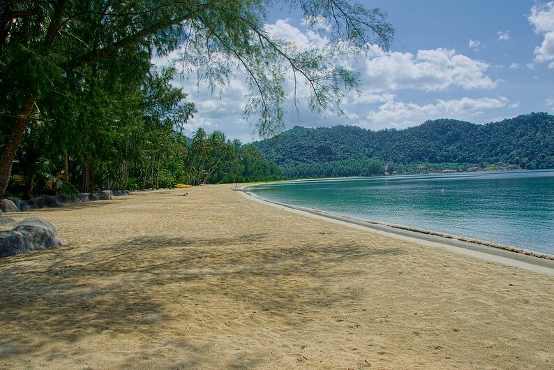 A sandy beach surrounded on the left by trees and on the right by clear blue water with mountains in the distance.