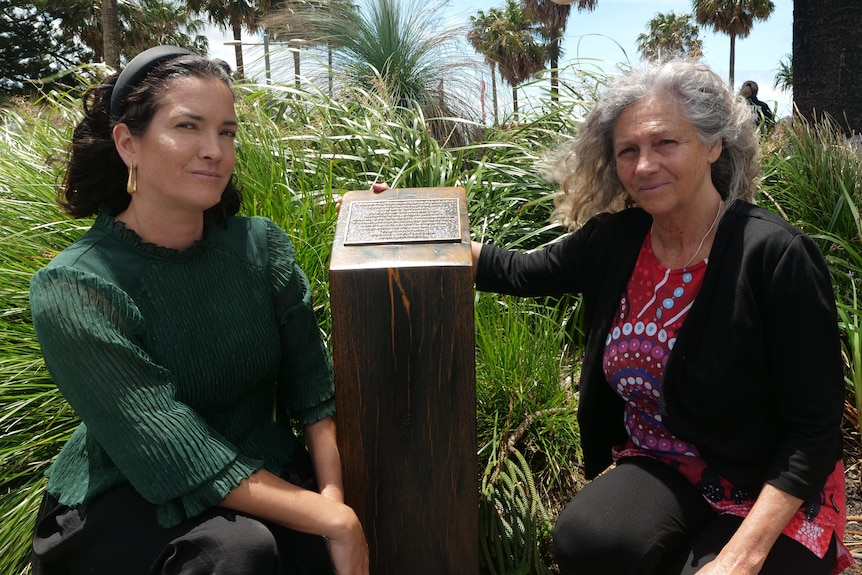 Mother and daughter sit beside a small plaque in front of a fenced garden.