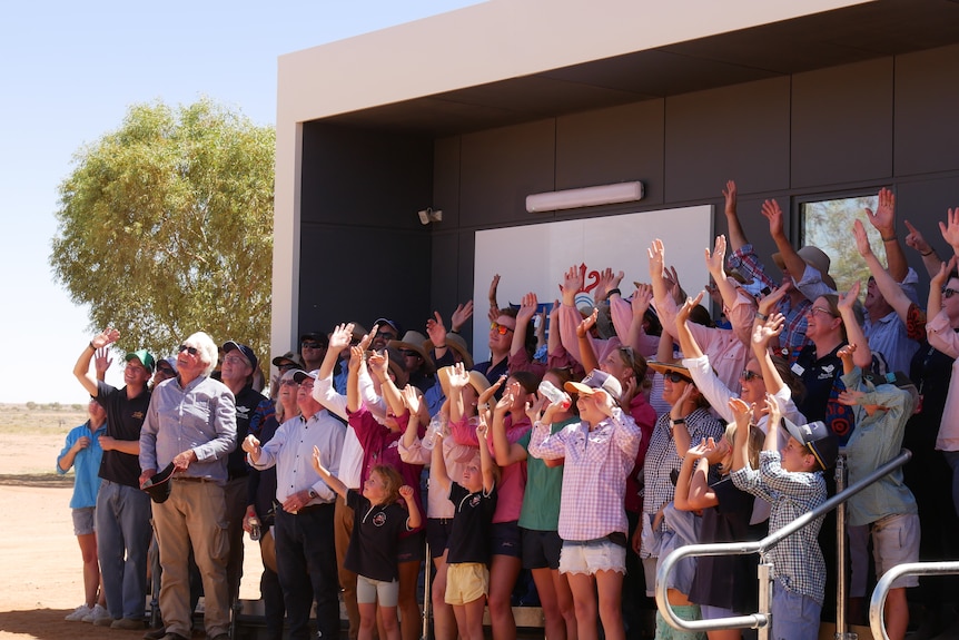 A group  of about 40 people stand in front of a building with their hands up waving at a drone. 