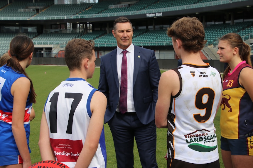 A man in a suit speaks to young football players in a sporting stadium.