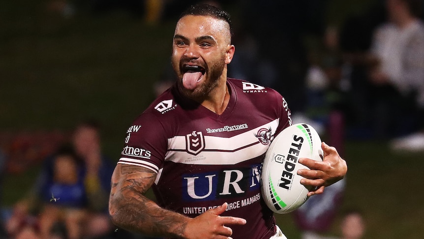 A Manly Sea Eagles NRL player pokes his tongue out as he runs with the ball in his left hand.