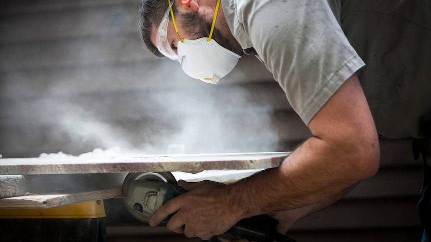 A man in a safety mask and goggles works on a countertop with a circular saw, surrounded by dust.