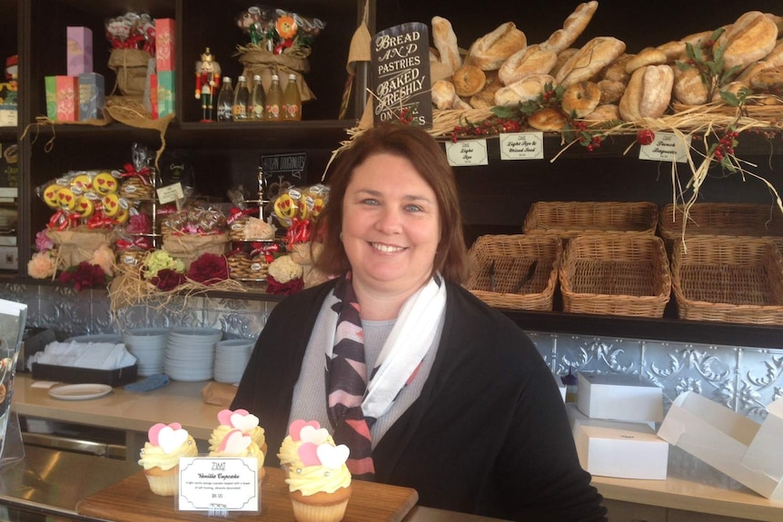 Woman stands in bakery with bread rolls behind her.