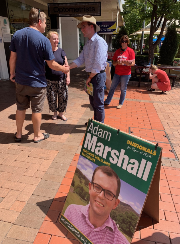 Sandwich board shows image of politcal candidate in foreground while candidate shakes hand with public in background