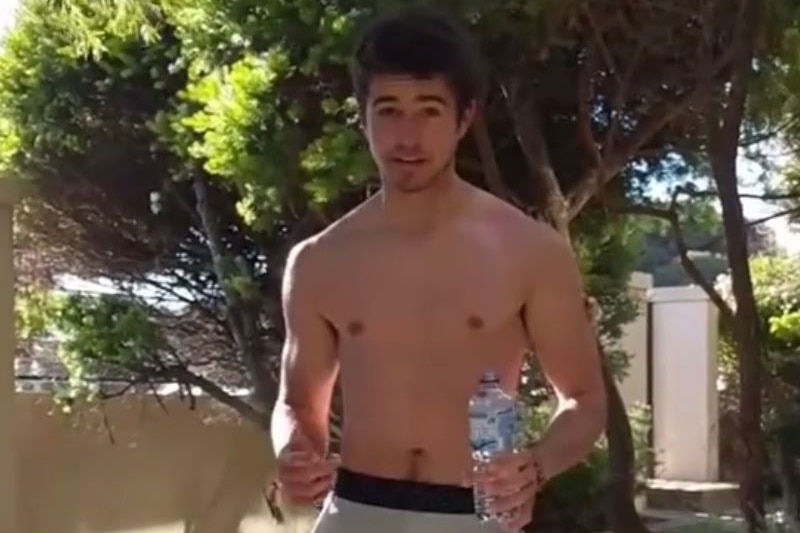 A young man in shorts and a bare chest holds a water bottle standing in front of a tree.