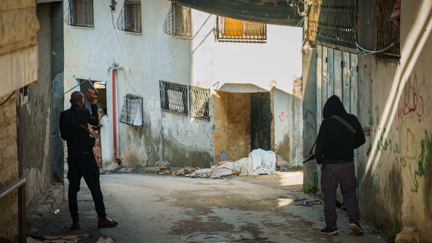 Two men dressed in black jumpers and carrying M16 rifles walk down an empty street.