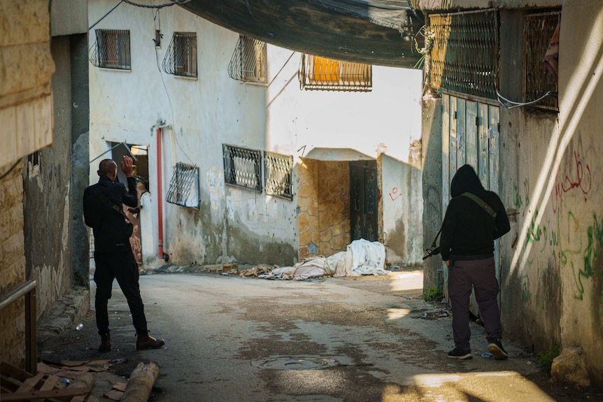 Two men dressed in black jumpers and carrying M16 rifles walk down an empty street.