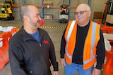 A bald man wearing a bright orange vests laughs with another man wearing a back jumper, they're in a warehouse.