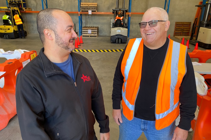 A bald man wearing a bright orange vests laughs with another man wearing a back jumper, they're in a warehouse.