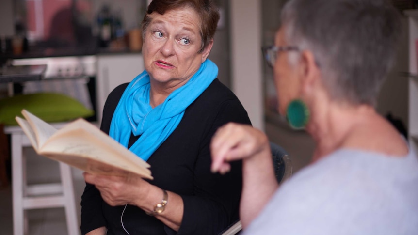 A older woman wearing a blue scarf is holding a book while giving a stern side-long glance to another woman