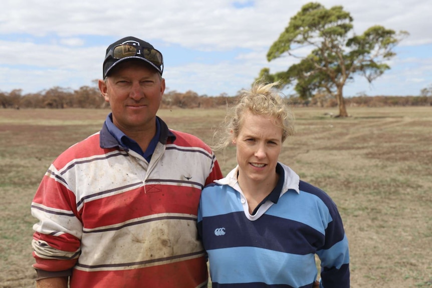 A man in a red striped rugby jumper and a cap on, stands with his arm around a woman in a blue striped rugby jumper in a paddock