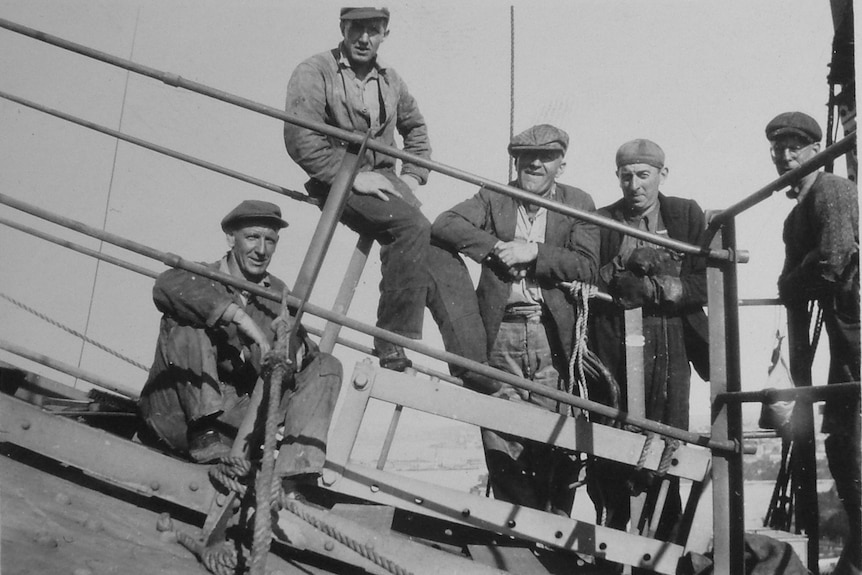 A black and white photo of five men with caps perched on a section of the bridge