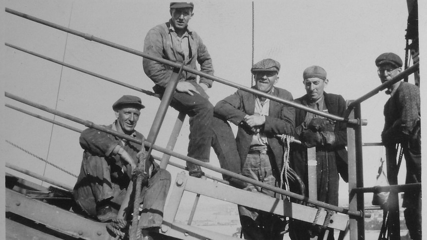 A black and white photo of five men with caps perched on a section of the bridge