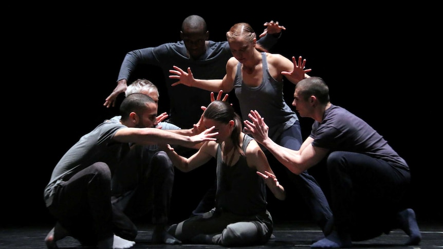 Five dancers with their arms up in a threatening manner, hover around a central dancer, sitting on the floor.