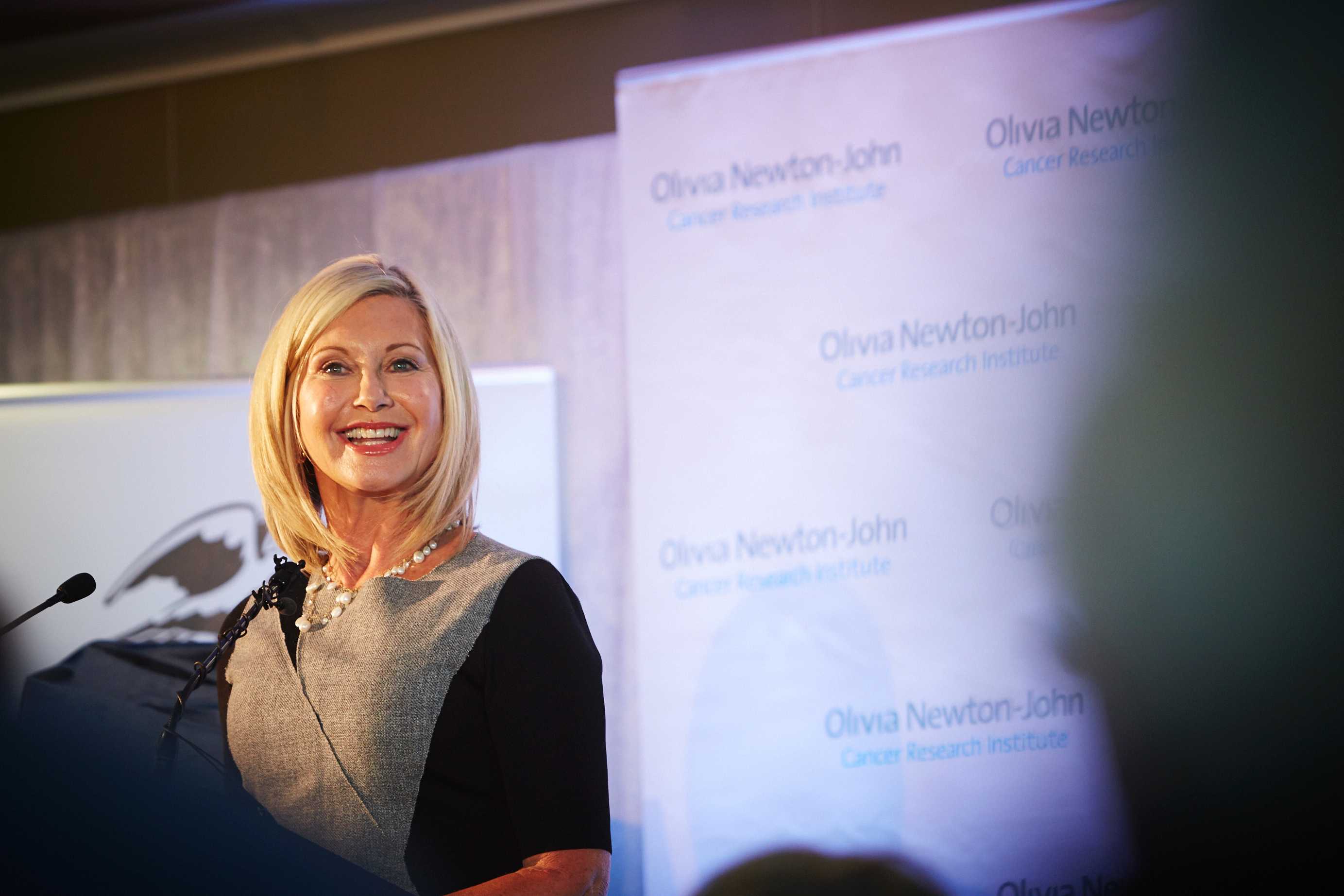 Olivia Newton-John smiles as she speaks to a podium microphone at the opening of a cancer research institute