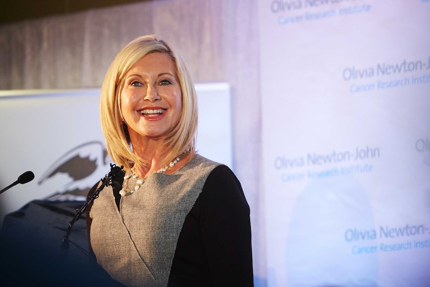 Olivia Newton-John smiles as she speaks to a podium microphone at the opening of a cancer research institute