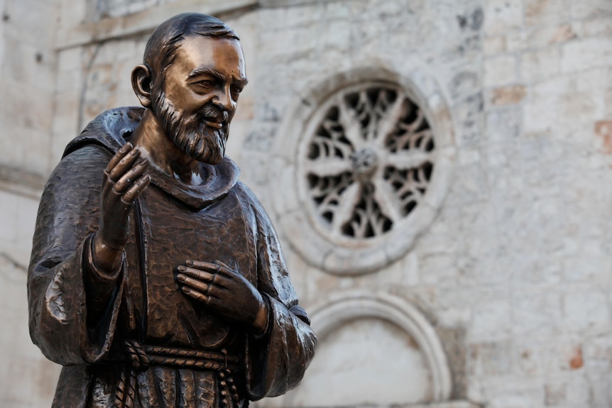 A bronze statue of Padre Pio, wearing hooded cloak and raising his right hand, smiling, with view of a cathedral in background.